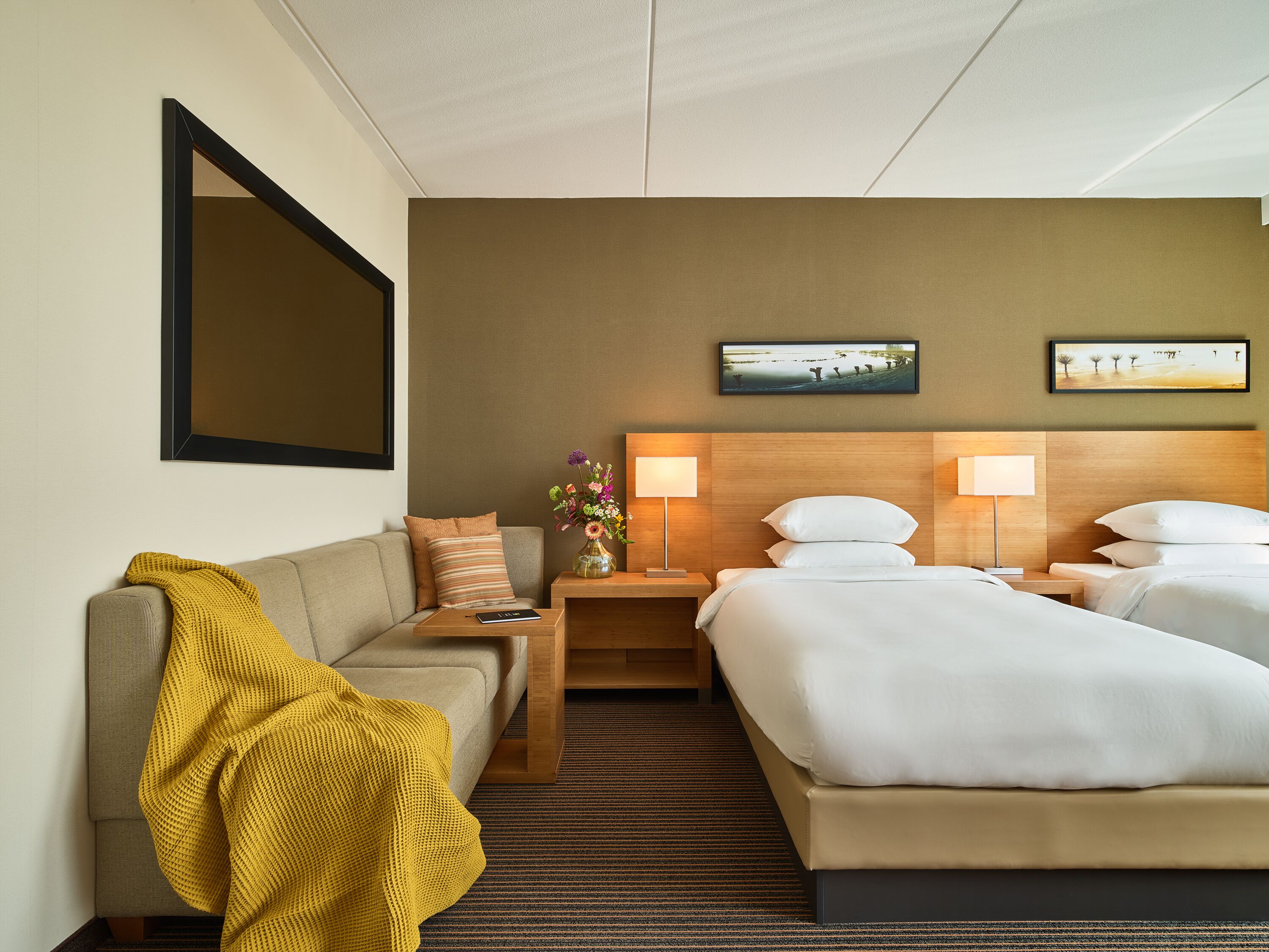 A hotel room with a sofa, two beds, a nightstand, a lamp, and a painting on the wall.