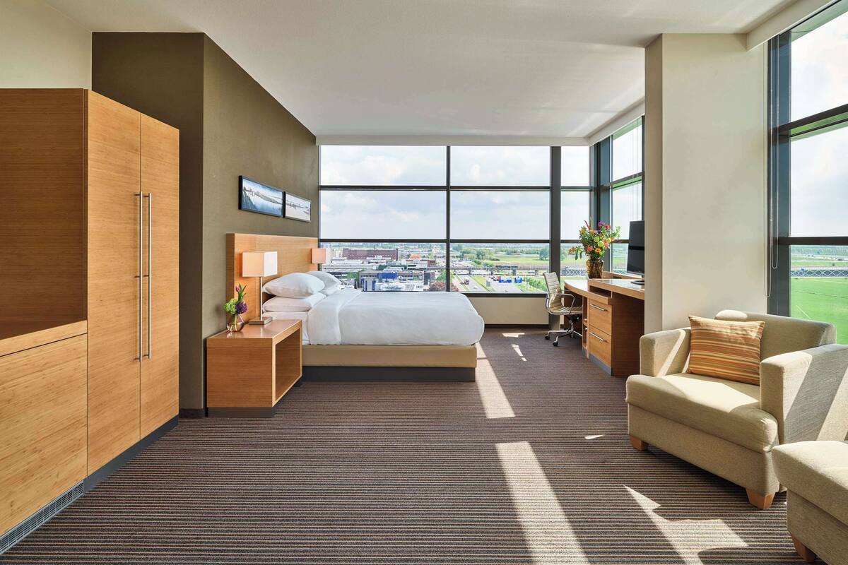 Une chambre d’hôtel moderne, dotée d’un grand lit, d’un bureau, d’une chaise et d’une armoire. La chambre dispose de grandes fenêtres offrant une vue sur la ville.