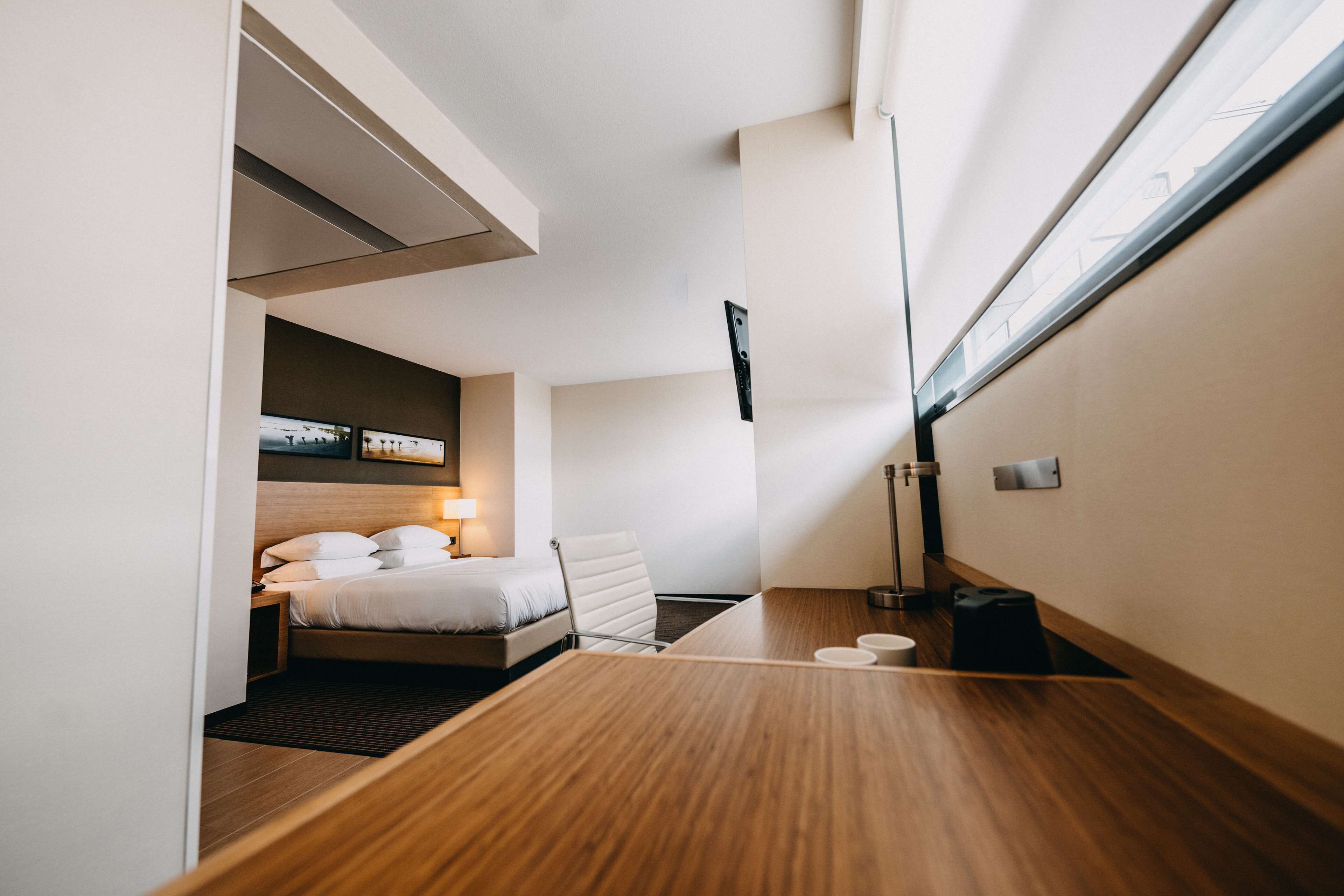 A modern hotel room with a bed, bedside table, lamp, and a view of the city through a large window.