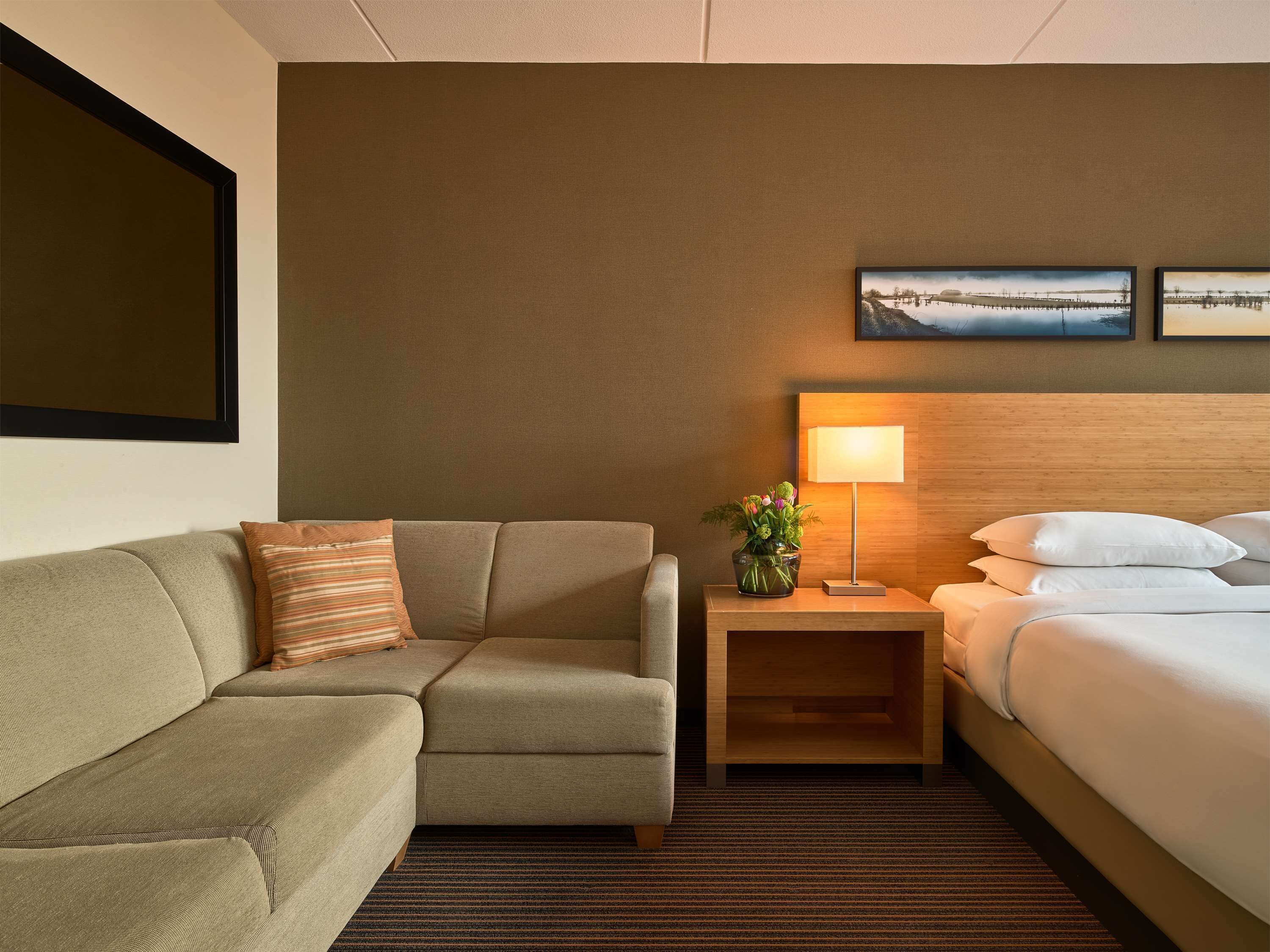 Habitación de hotel con sofá, mesita de noche con lámpara, cama y dos cuadros en la pared.