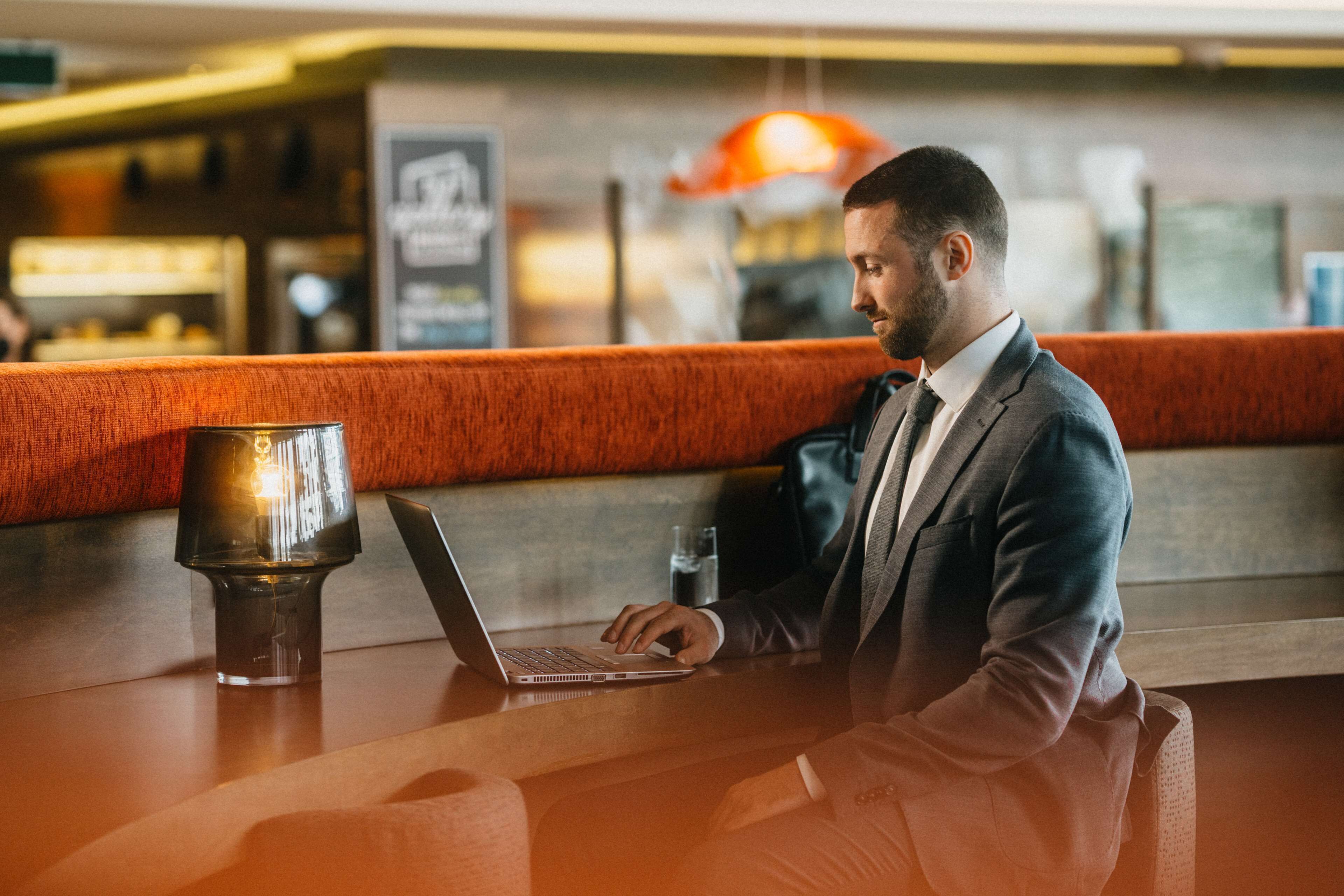 A man in a suit working on a laptop at a bar with a glass of water and a lamp.