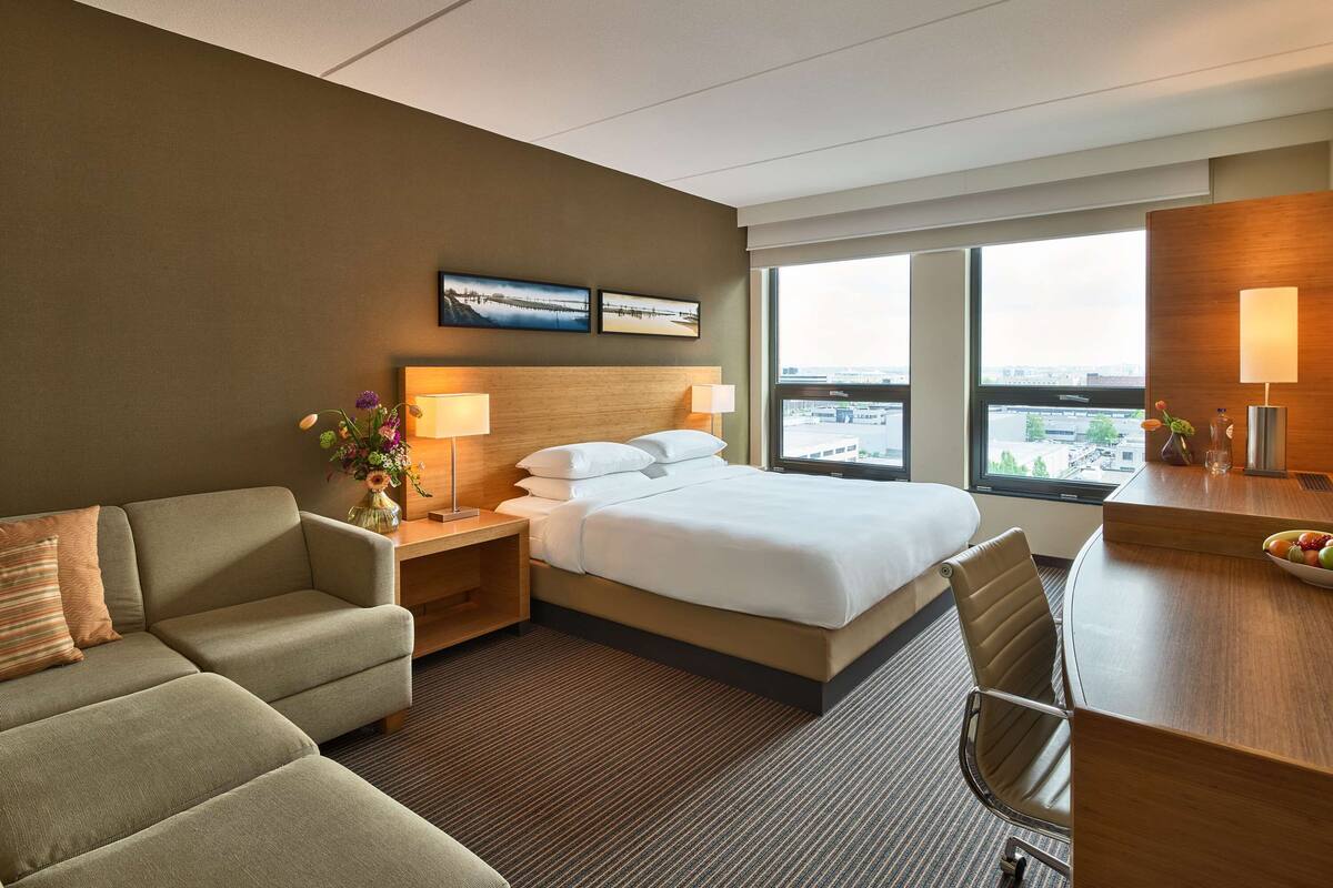 Une chambre d’hôtel équipée d’un lit, d’un canapé, d’un bureau et d’une grande fenêtre donnant sur la ville.