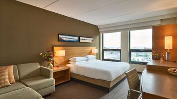 A hotel room with a bed, a sofa, a desk, and a large window offering a city view.