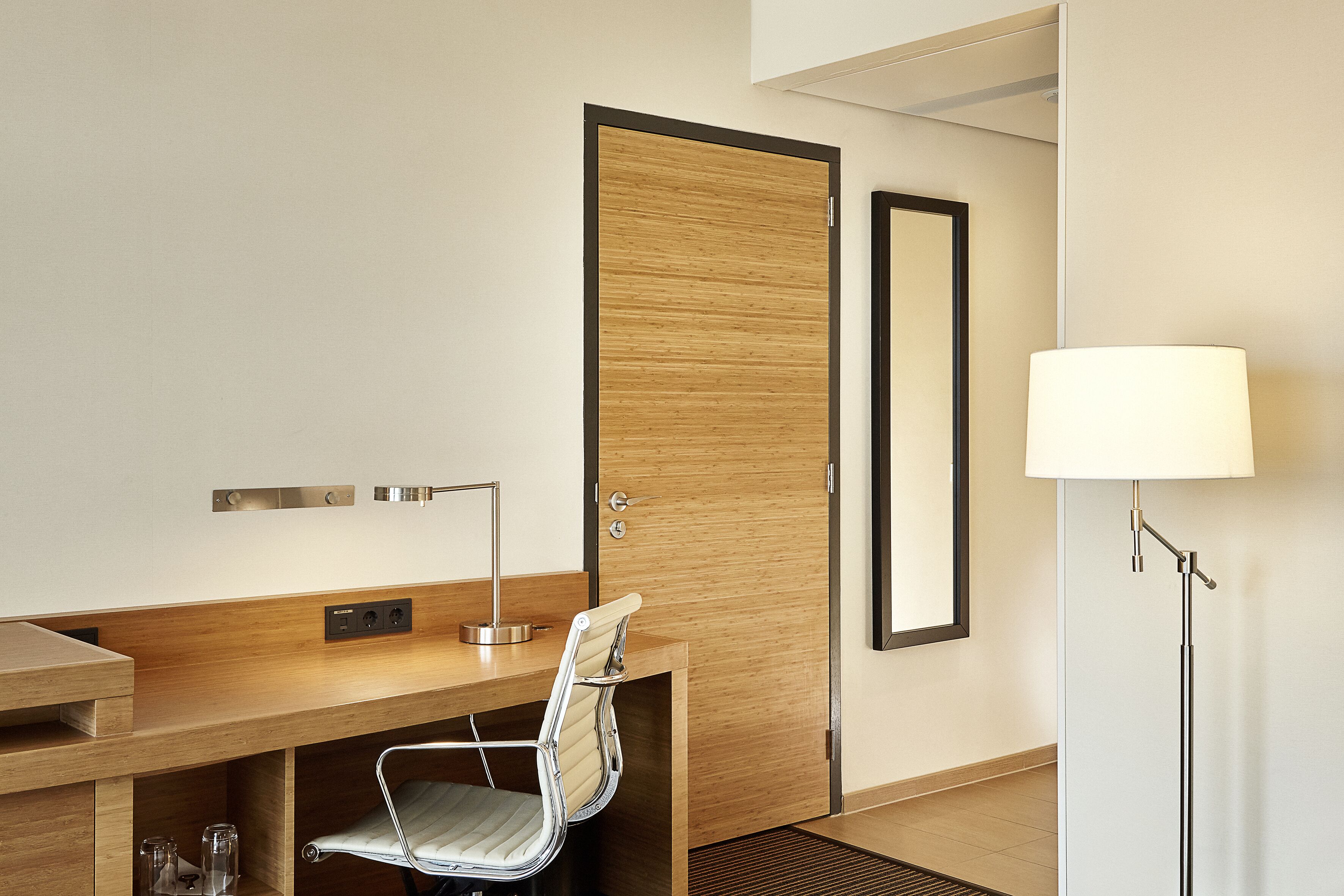 Quarto de hotel com uma escrivaninha de madeira, uma cadeira, um abajur, um espelho e uma porta.