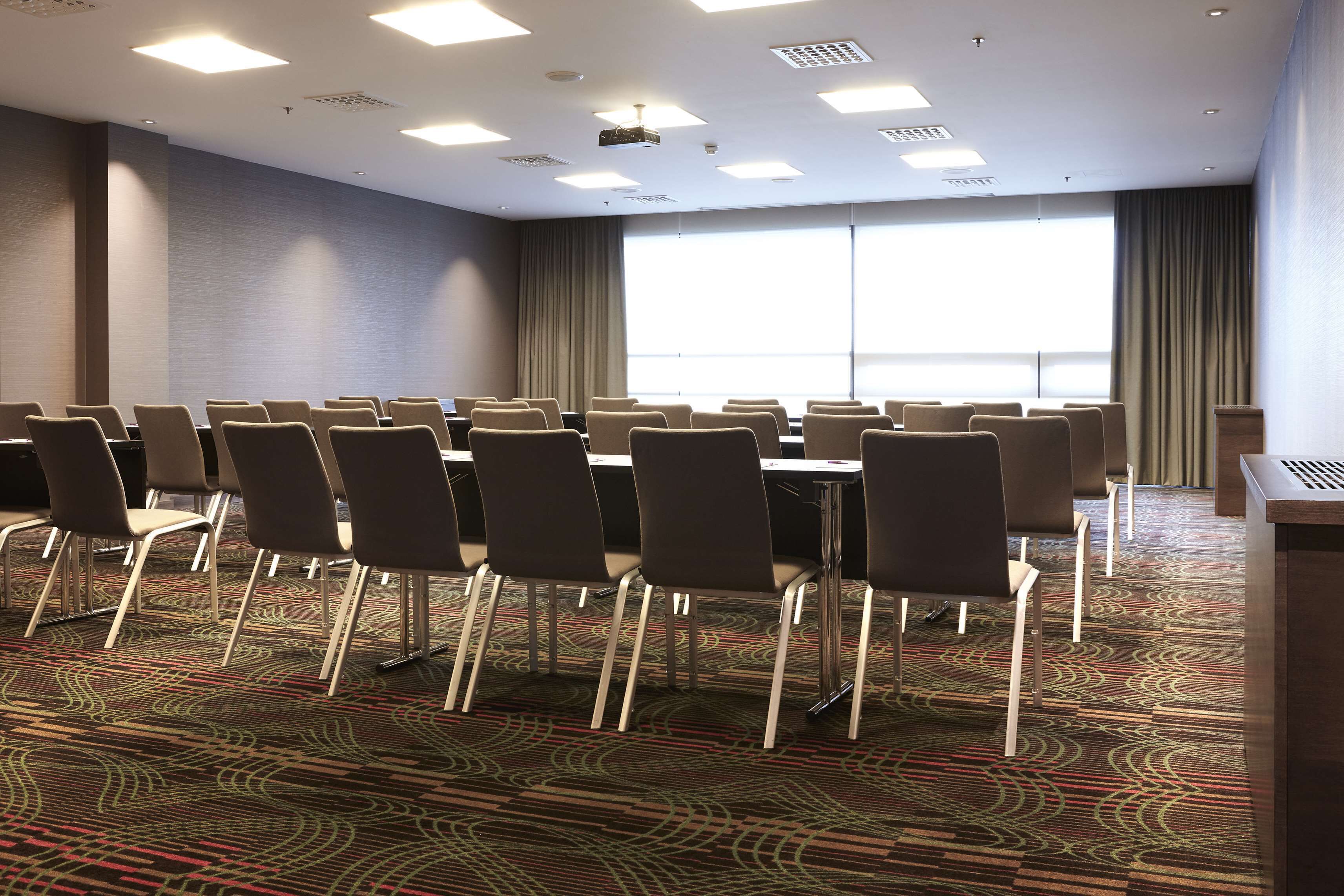 Un salón de conferencias con filas de sillas, un proyector y un ventanal con cortinas.