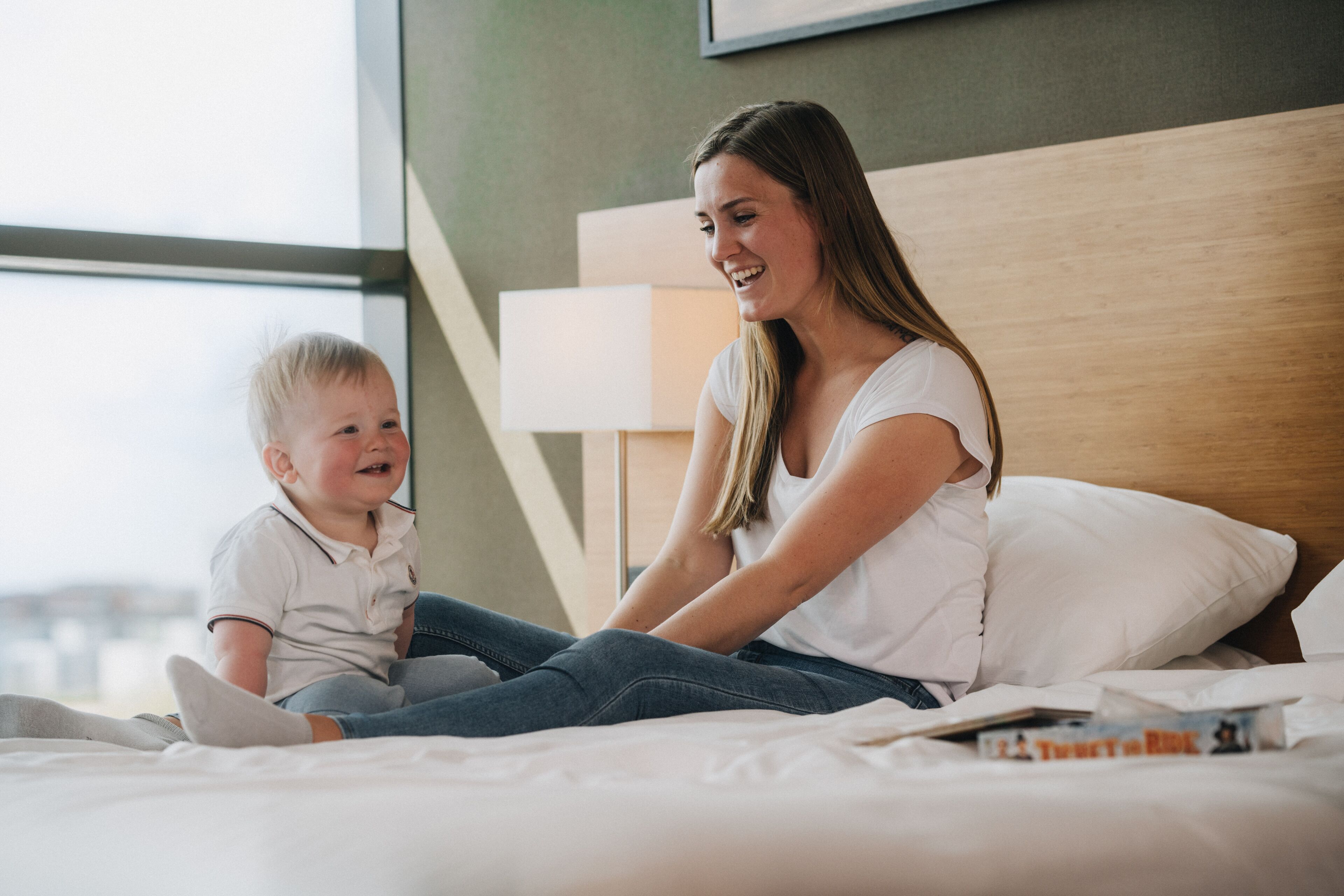 Una mujer y un niño sentados en una cama en una habitación con un ventanal grande y un cabecero de madera.
