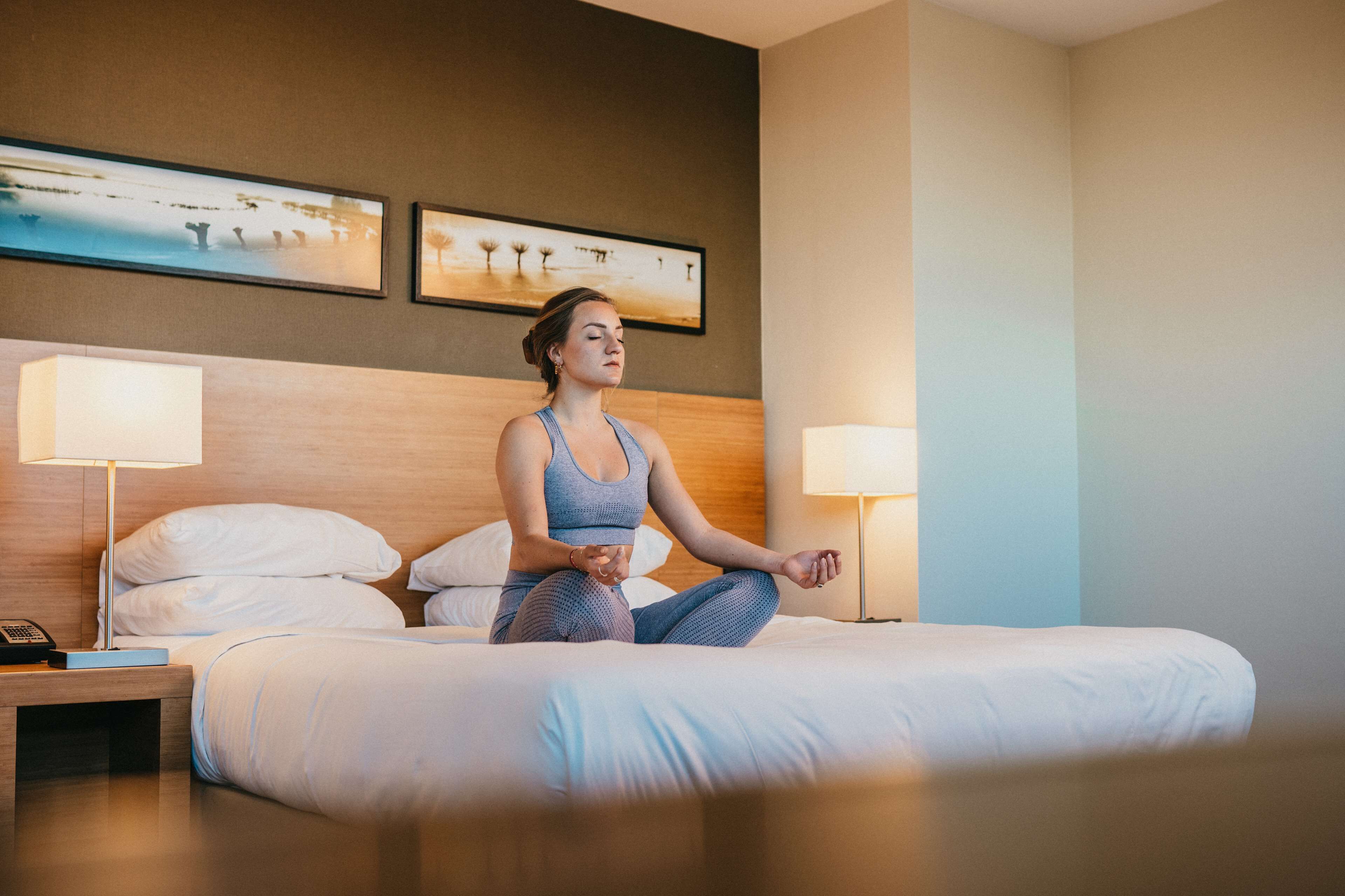 Quarto de hotel com uma cama, duas luminárias de cabeceira, mesa de cabeceira com telefone e duas fotografias emolduradas na parede.
