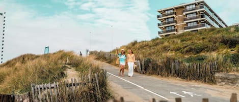 Un sendero conduce a un edificio con varios balcones, rodeado de dunas cubiertas de hierba.