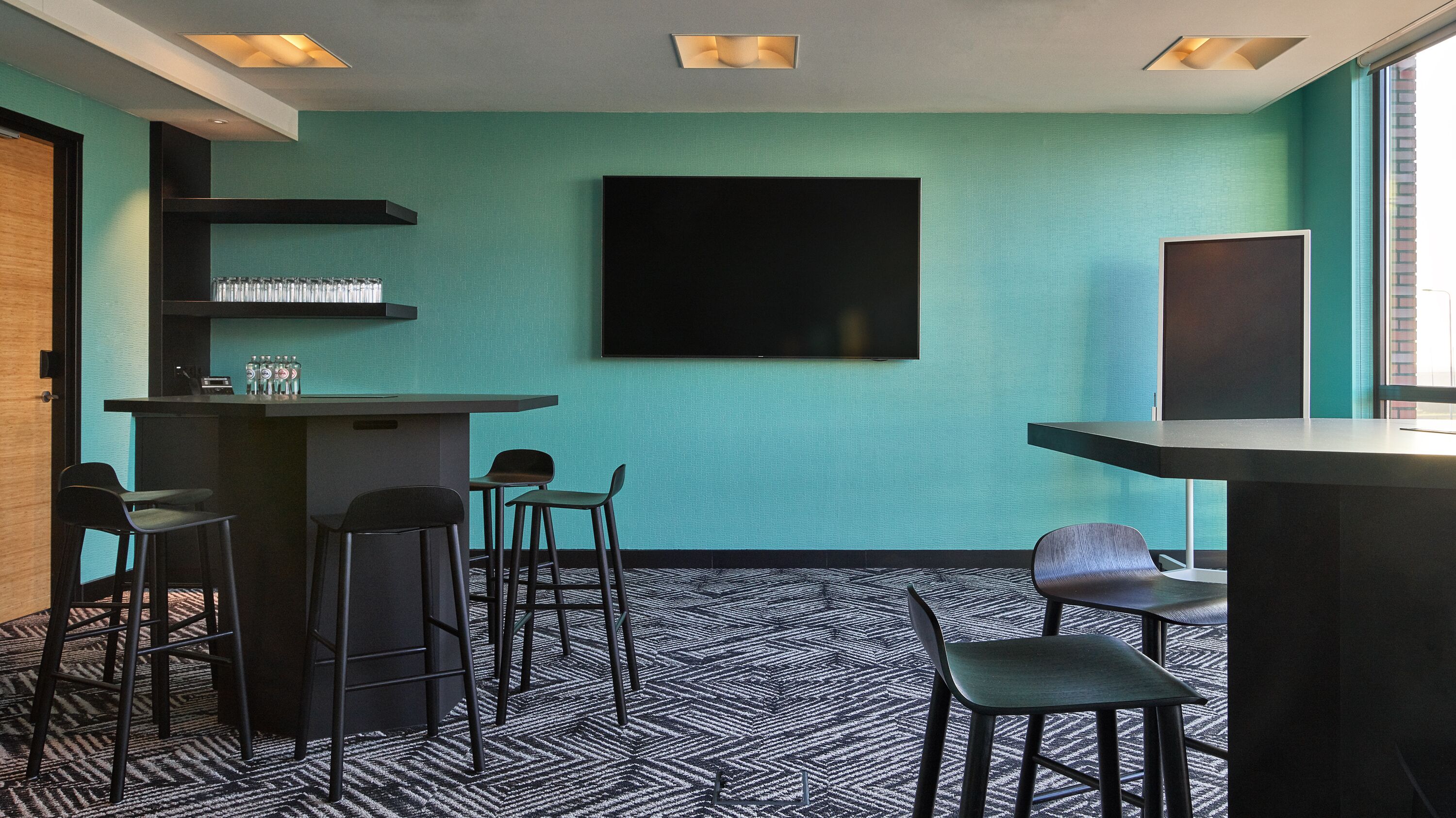 A modern bar area with a teal wall, black bar stools, and a patterned carpet.