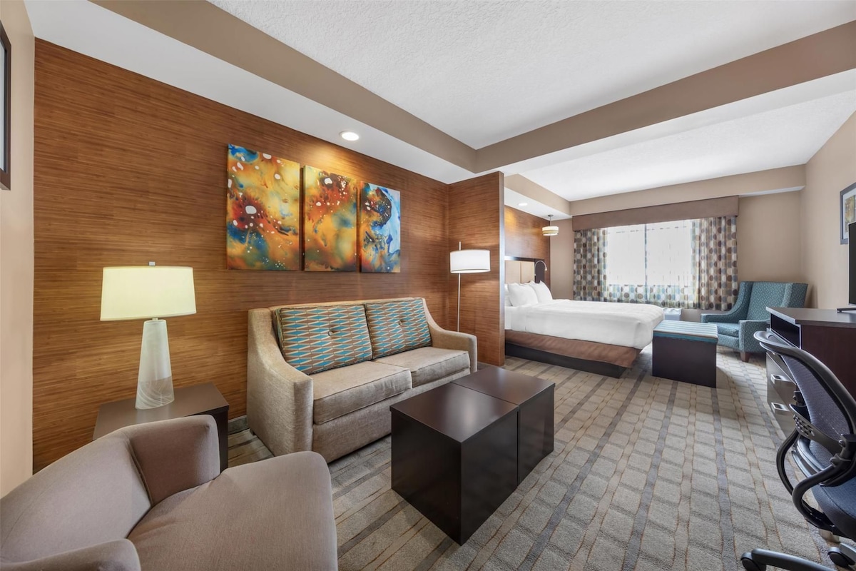 A hotel room with a bed, sofa, armchair, coffee table, and artwork on the wall.