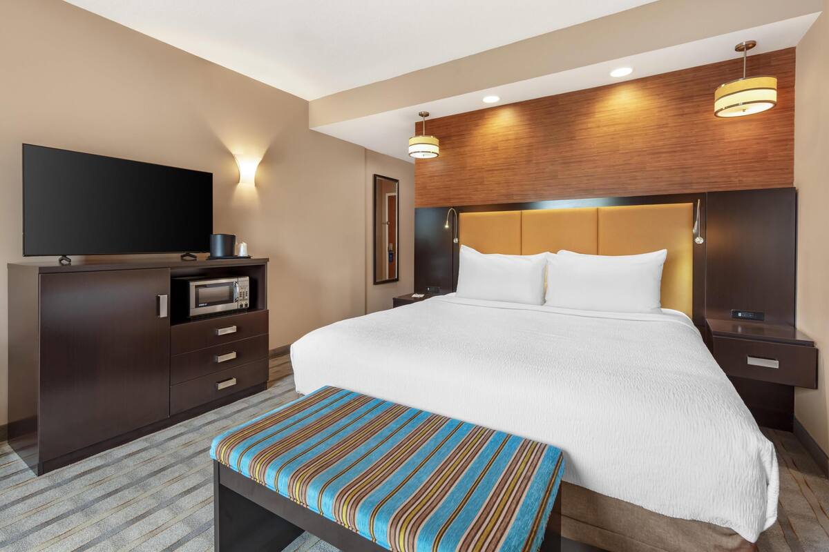 Ein Hotelzimmer mit einem großen Bett, einem Flachbildfernseher, einem hölzernen Kopfteil und einer Bank mit gestreifter Sitzkissen.