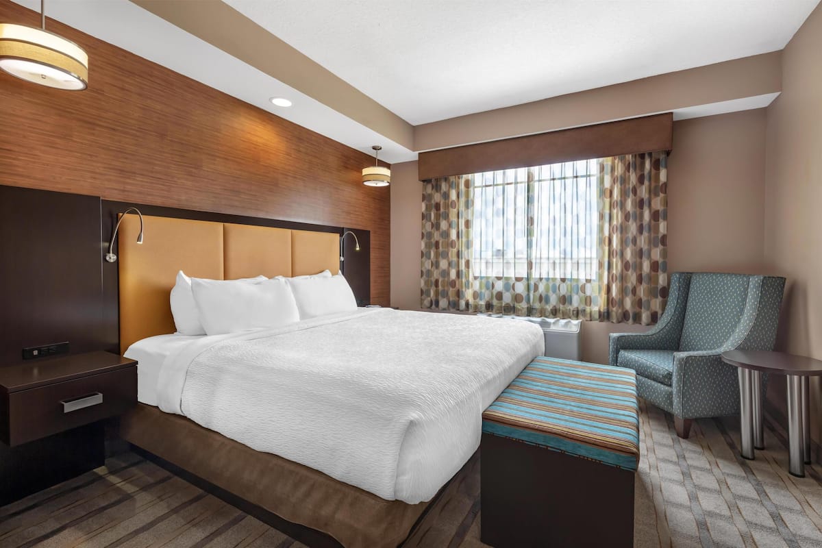 Ein Hotelzimmer mit einem großen Bett, einem Nachttisch, einem Sessel, einem kleinen Tisch und einem Fenster mit Vorhängen.