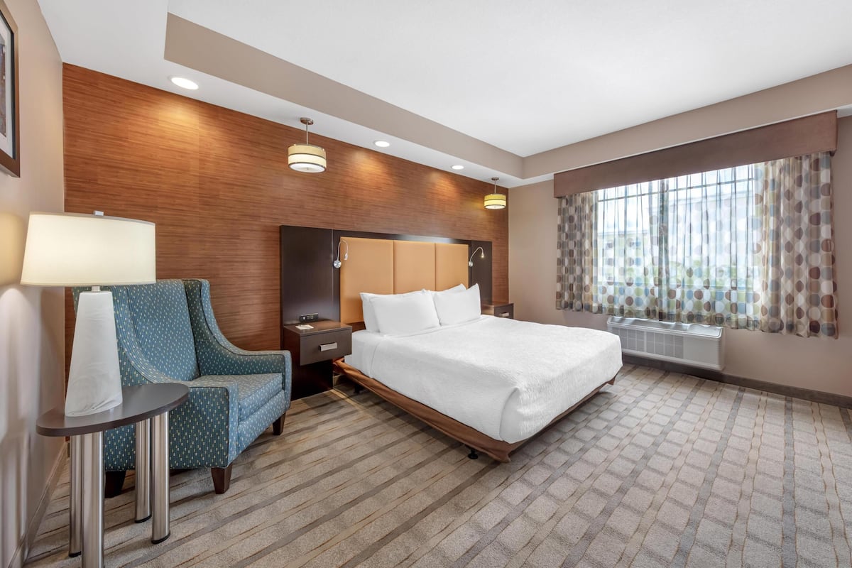 Ein Hotelzimmer mit einem Bett, einem Sessel, einem Nachttisch, einer Lampe, einem Fenster mit Vorhängen und einer Wand mit Holzpaneelen.