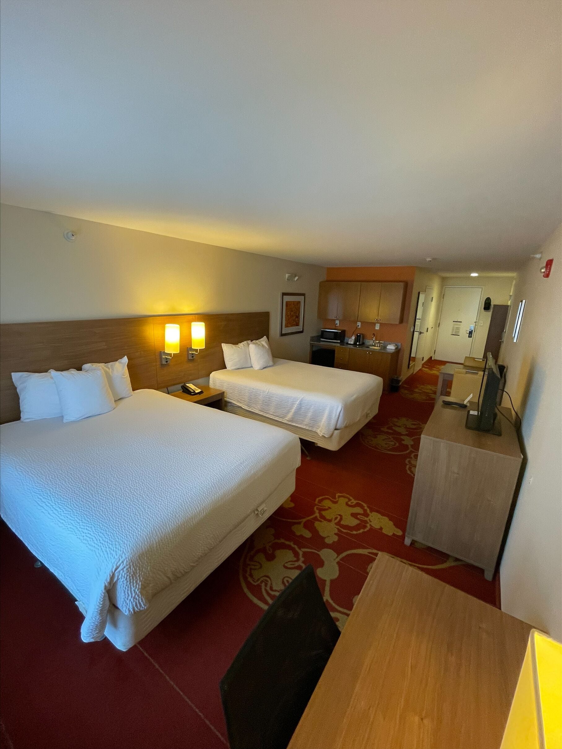 A hotel room with two beds, a desk, a chair, a microwave, and a small kitchenette.