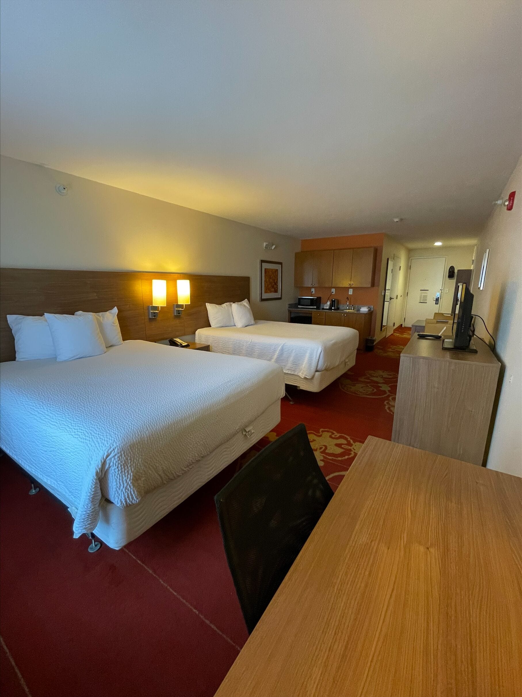 Habitación de hotel con dos camas, una kitchenette pequeña, un escritorio y un televisor.