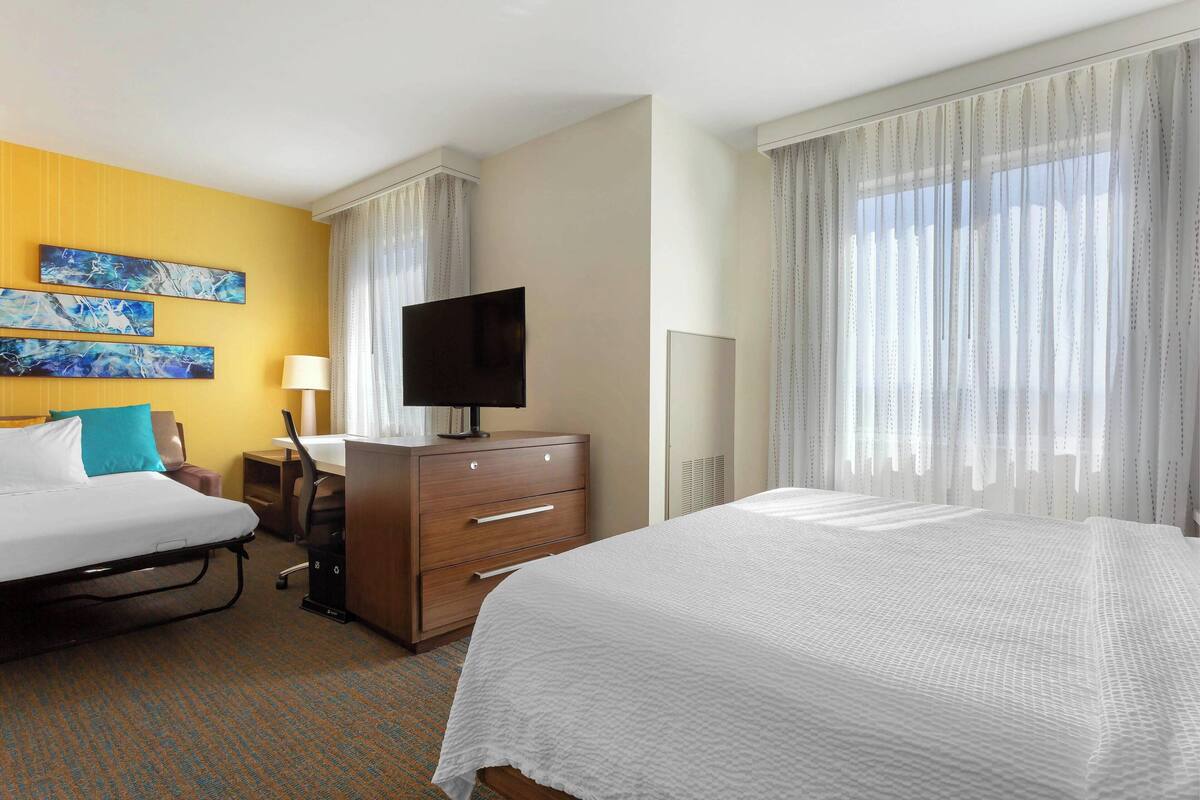 A hotel room with two beds, a desk, a TV, and artwork on the wall.