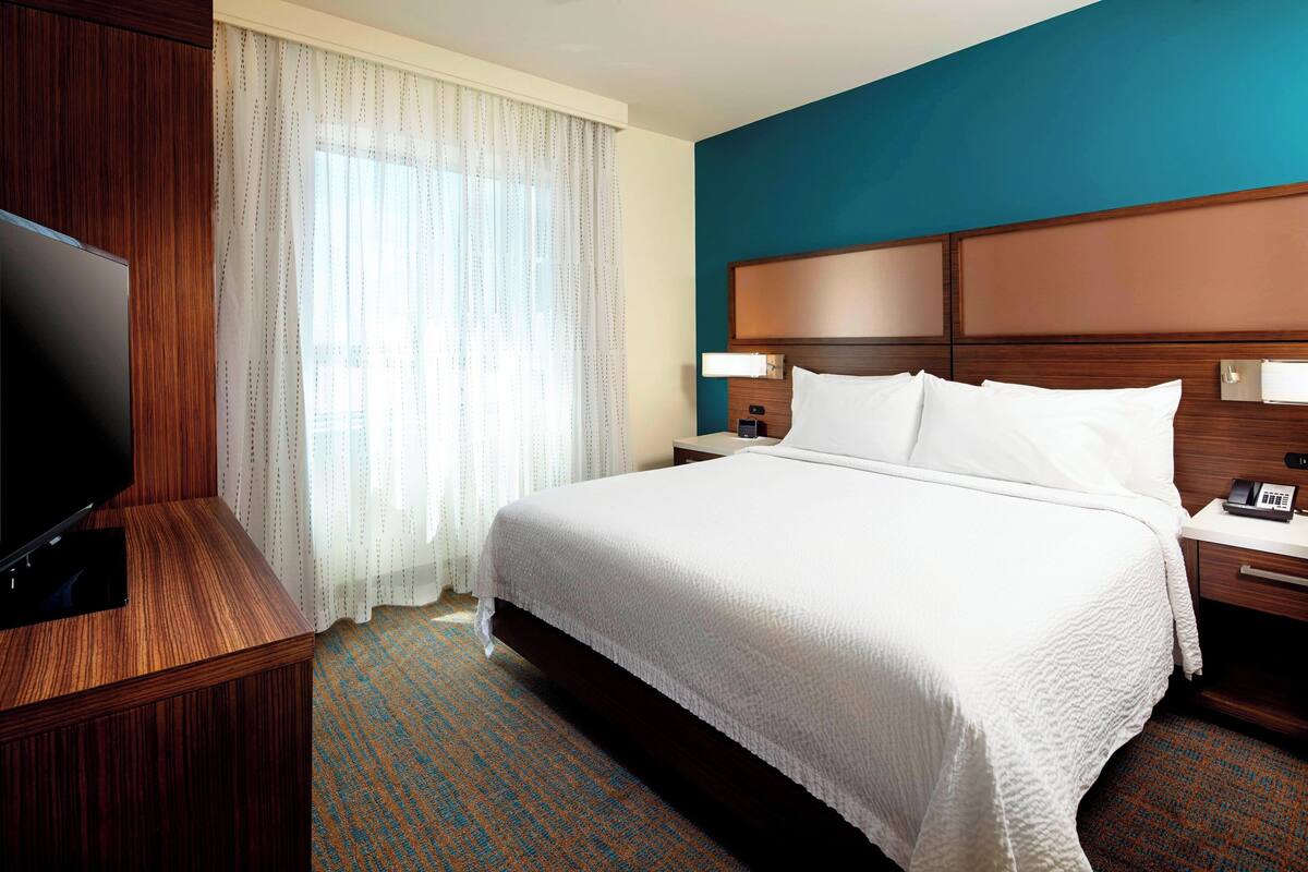 A hotel room with a large bed, a TV, a bedside table with a lamp, and a window with sheer curtains.
