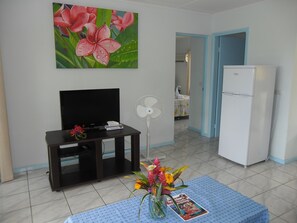 Un soggiorno con televisore, ventilatore, frigorifero e un tavolo con fiori e una rivista.