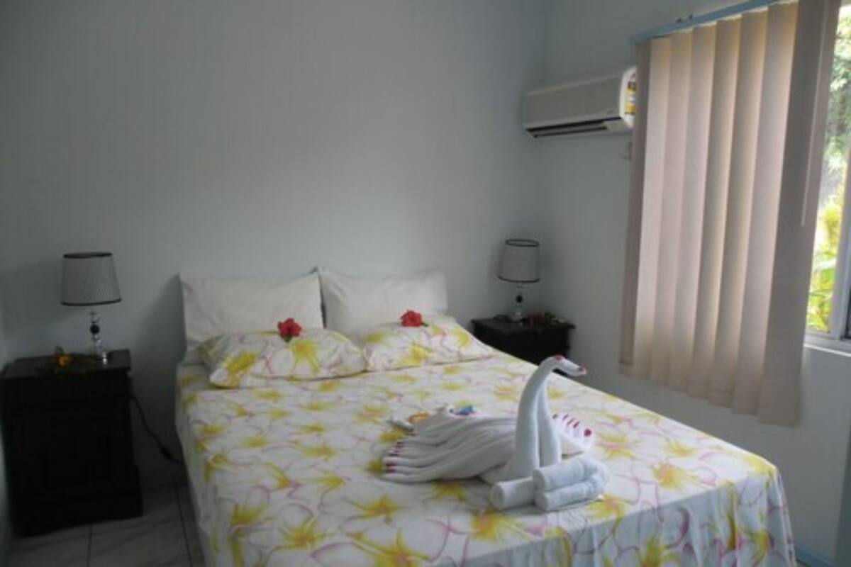 A hotel room with a bed, two bedside tables, an air conditioning unit, and a window with blinds.