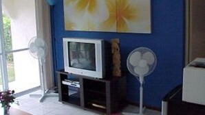 A room with a blue wall, a television on a stand, and two standing fans. - Quest Apartments (Port Vila)