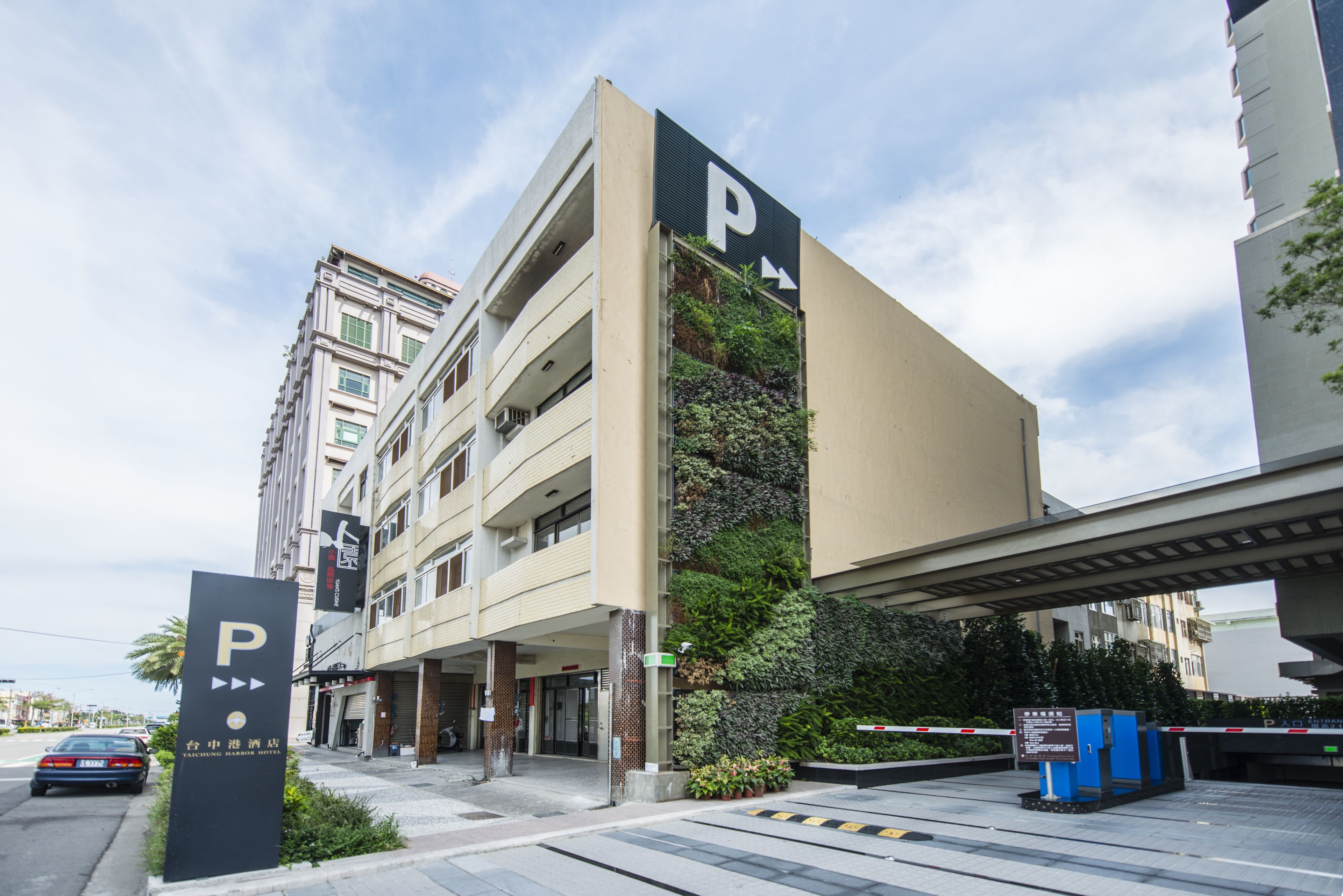A multi-story hotel with a parking entrance, a vertical garden on the facade, and a pedestrian overpass above.