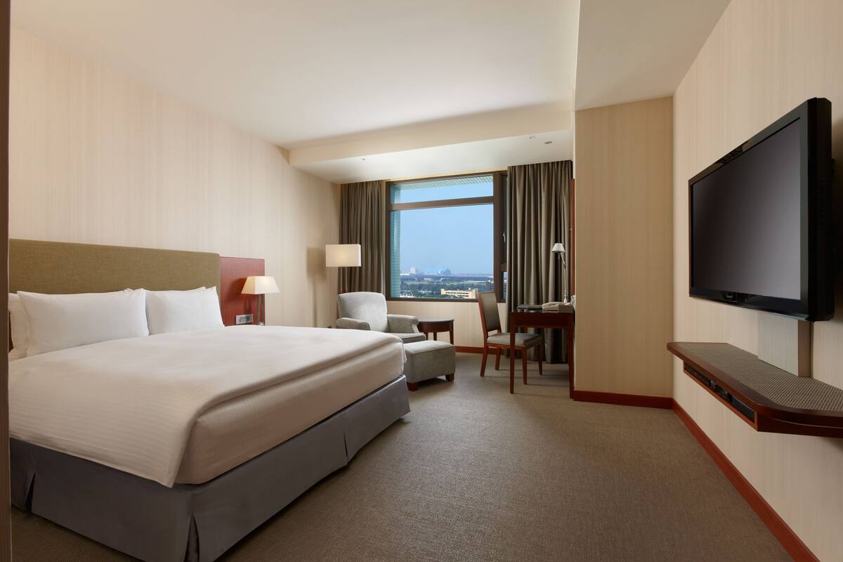 A hotel room with a large bed, a desk, a chair, a television, and a view of the city.