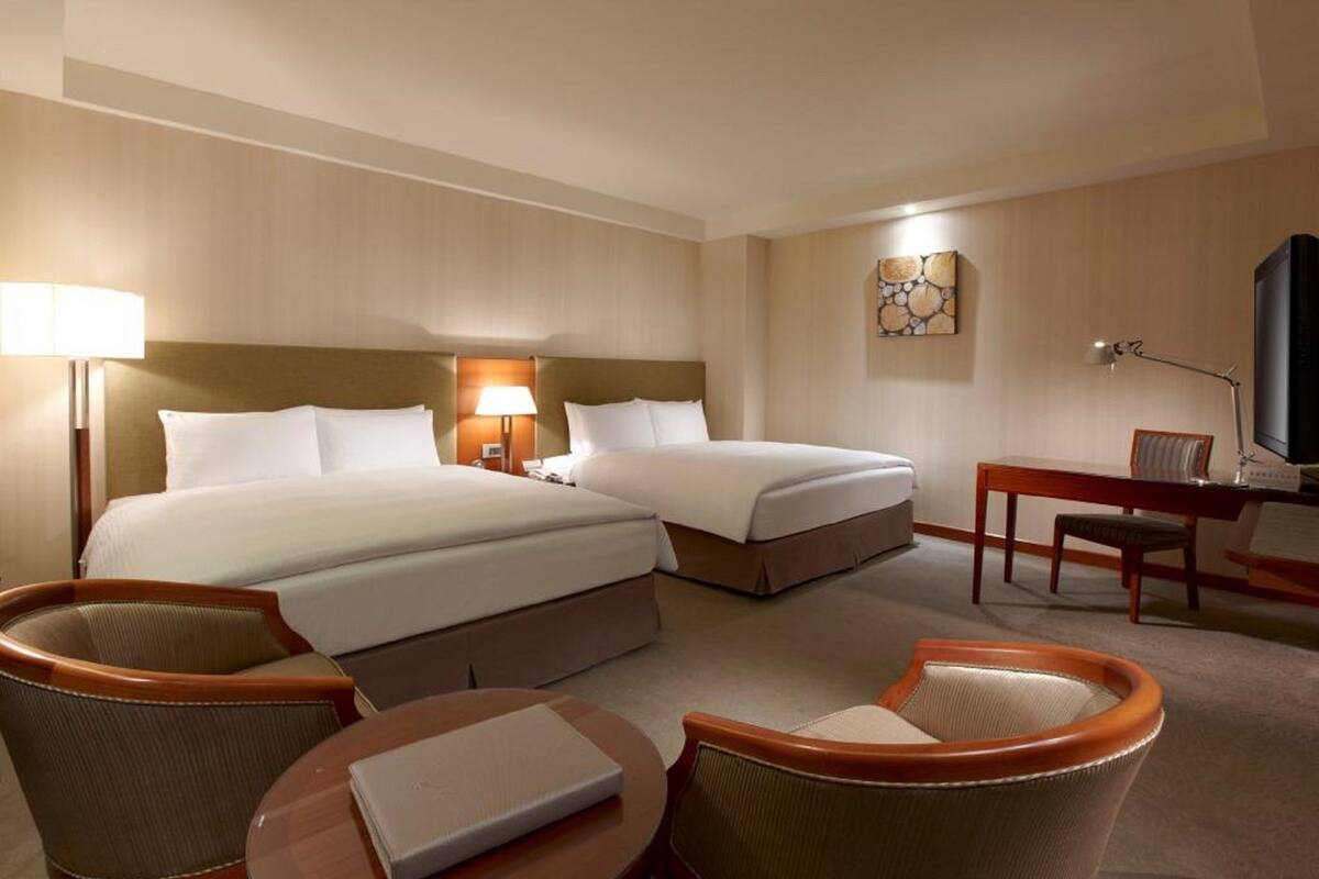A hotel room with two beds, a desk, a chair, and a lamp.