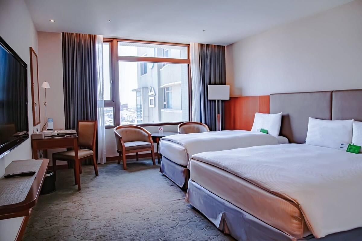 A hotel room with two beds, a desk, a chair, a TV, and a window with curtains.