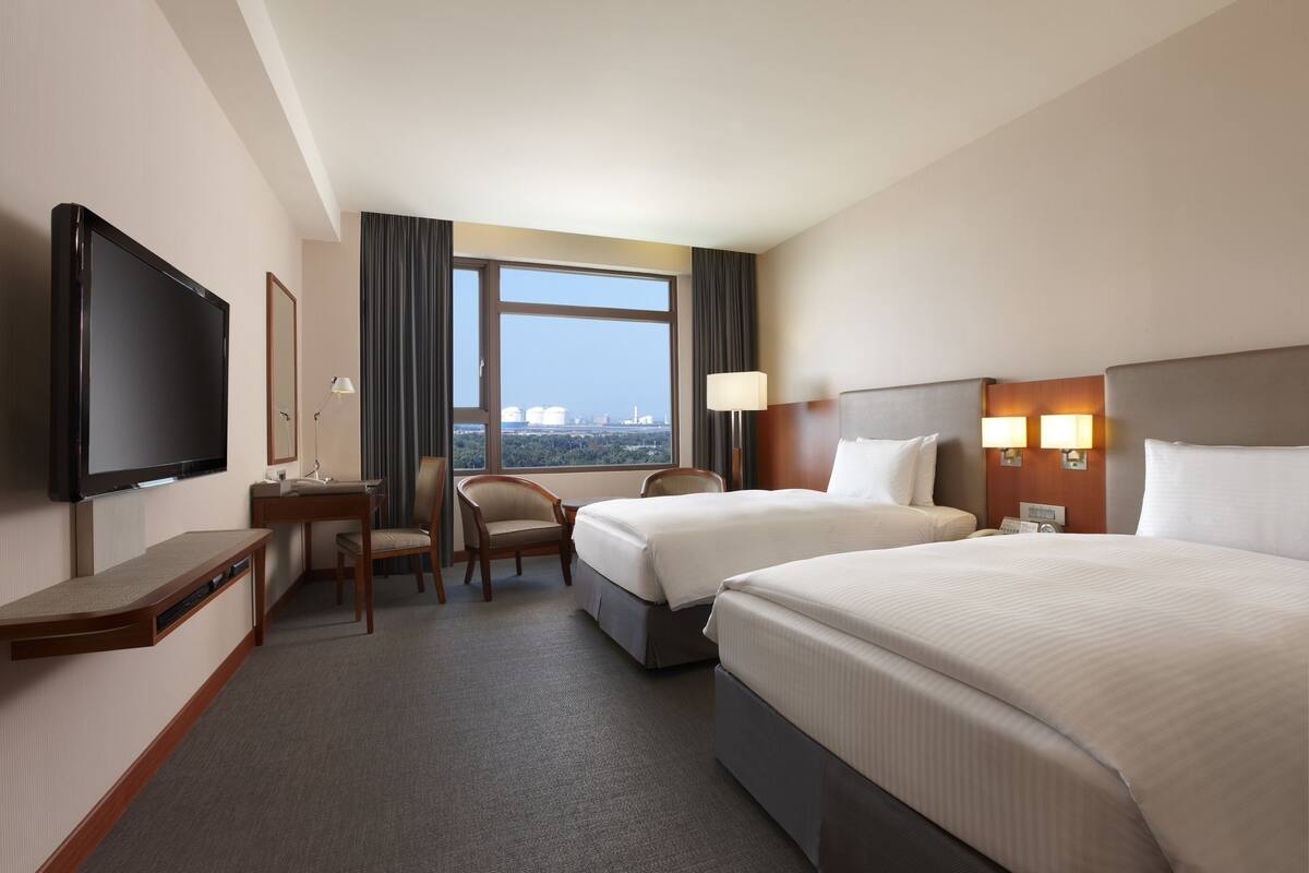 A hotel room with two beds, a desk, a chair, a TV, and a large window with a city view.