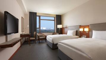 A hotel room with two beds, a desk, a chair, a TV, and a large window with a city view.