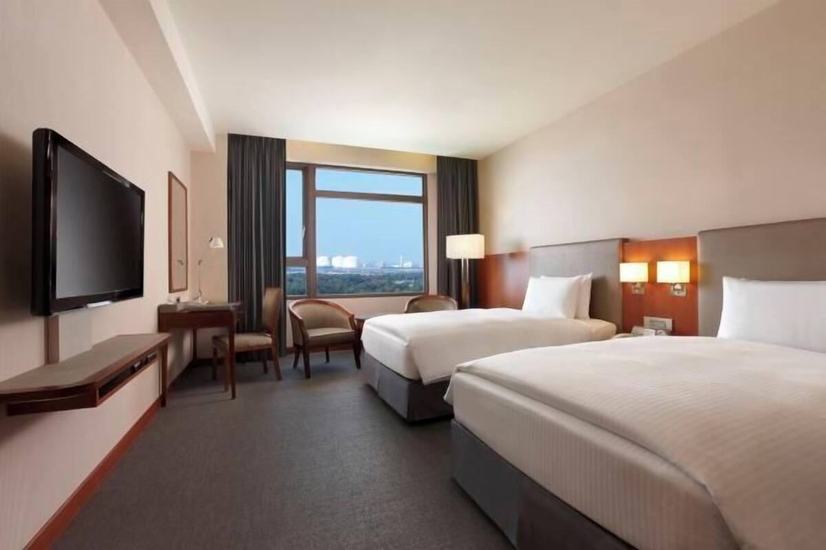 A hotel room with two beds, a desk, a chair, a TV, and a window with a city view.