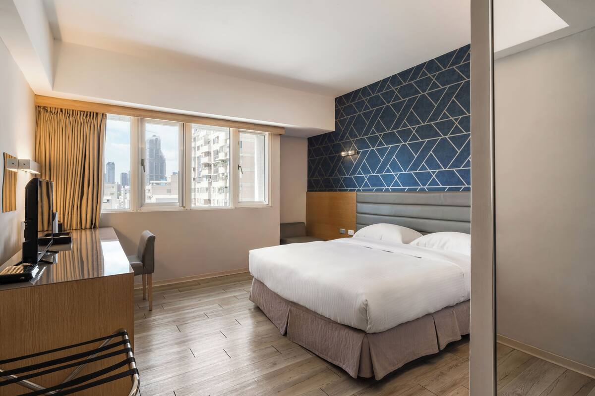 A hotel room with a bed, a desk, and a chair. There is a large window with a view of buildings.