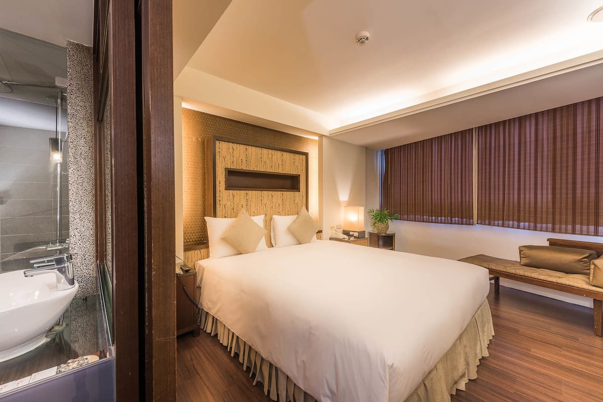 Habitación de hotel con una cama grande, cabecero de madera, mesita de noche con lámpara, un banco y vistas a la ciudad a través de cortinas.