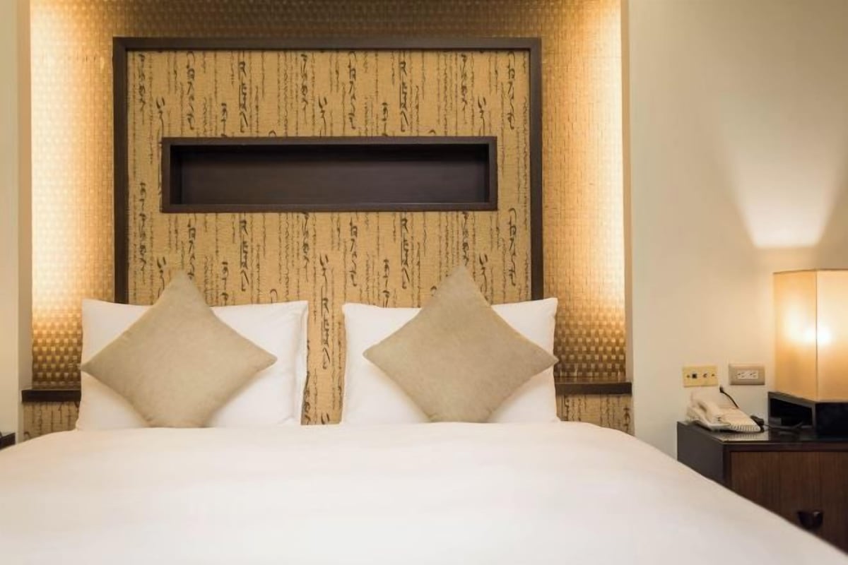 Quarto de hotel com uma cama grande, duas almofadas, uma mesa de cabeceira com abajur e uma parede com padrão texturizado.