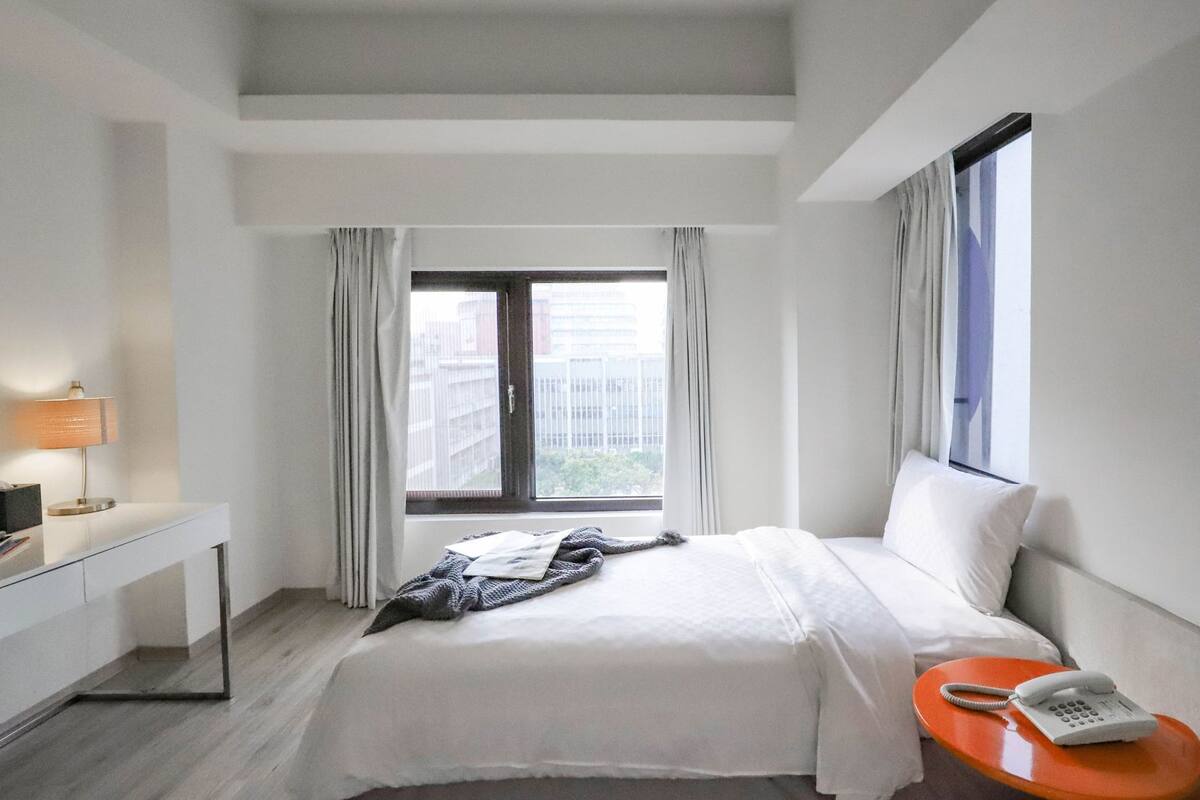 Een hotelkamer met een bed, een bureau, een telefoon en een raam met gordijnen.