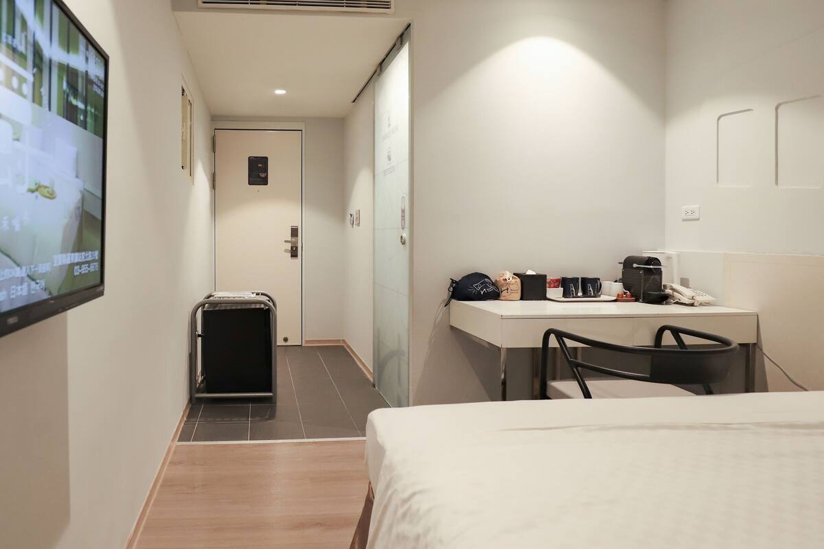 Een hotelkamer met een bed, een bureau met stoel, een aan de muur bevestigde televisie en een koffer op de vloer.