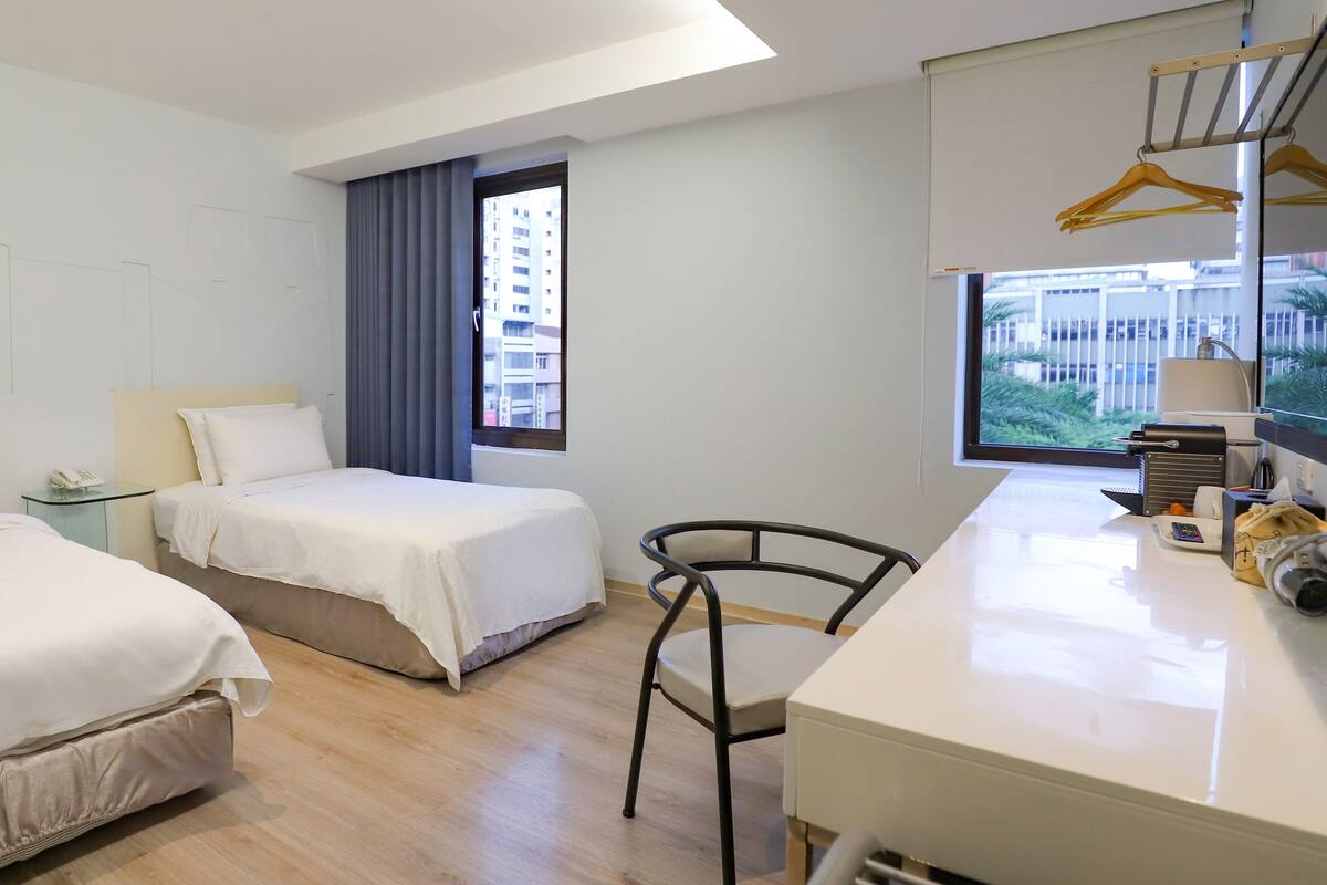 Een hotelkamer met twee bedden, een bureau, een stoel, een raam met gordijnen en uitzicht op gebouwen.