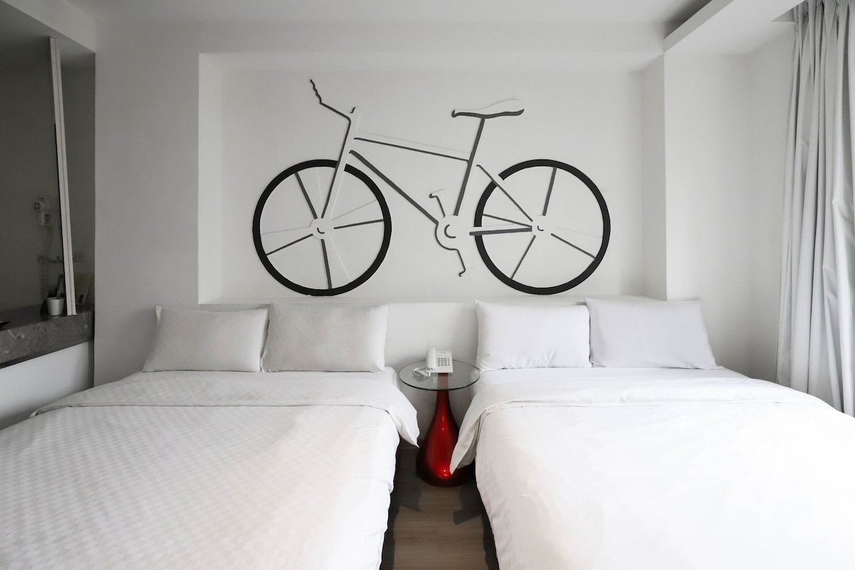 Ein Hotelzimmer mit zwei Betten, einem an der Wand befestigten Fahrradbild, einem kleinen Tisch mit Telefon und einer roten Vase.