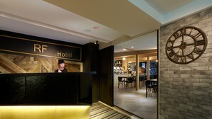 Zona de recepción de un hotel con un mostrador de madera, un reloj grande en la pared y una vista del comedor al fondo.