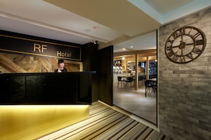 Een hotel receptie met een houten balie, een grote klok aan de muur, en een blik op de ontbijtruimte op de achtergrond.