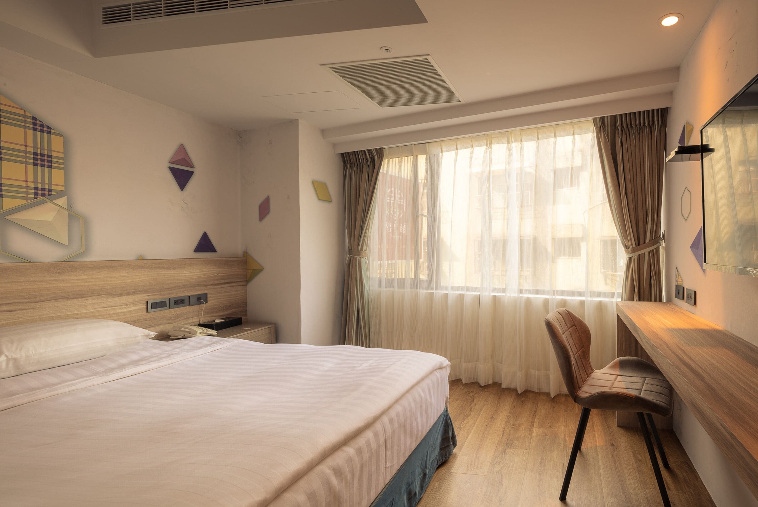 A hotel room with a bed, a desk, and a chair. There is a window with curtains, a wall clock, and a wall-mounted light fixture.