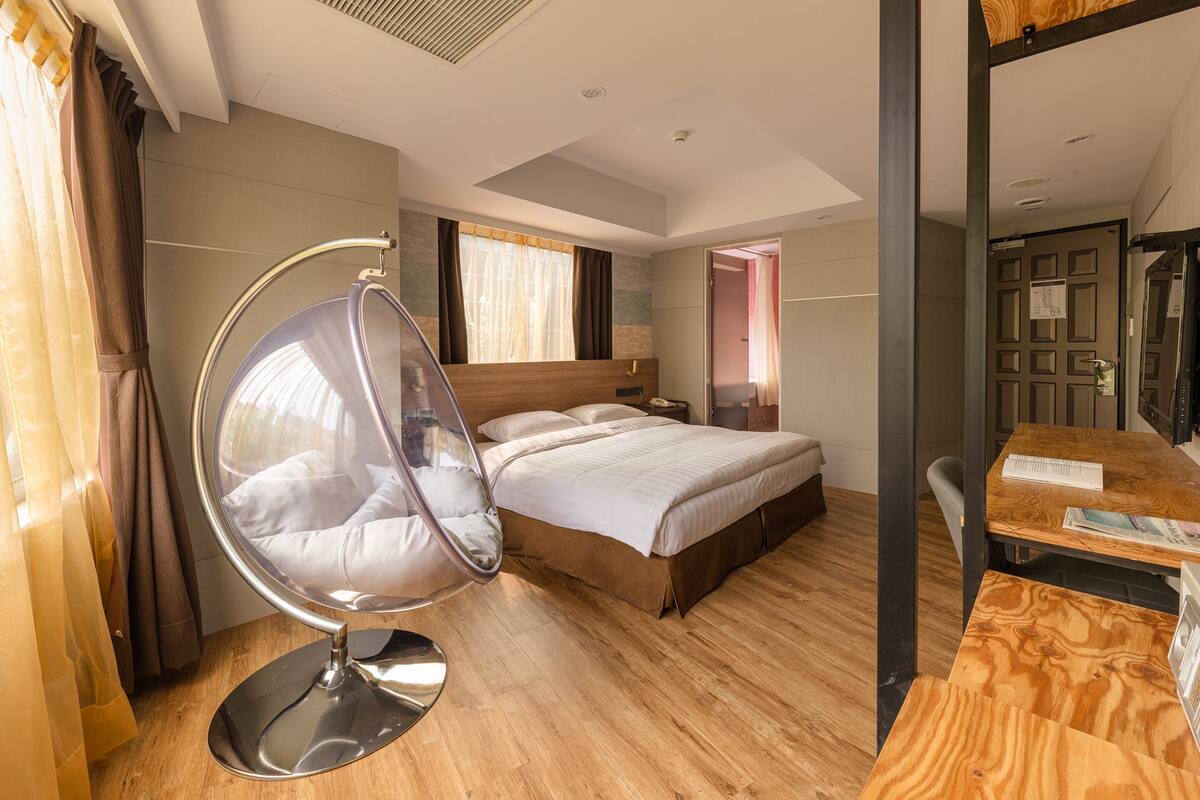 A modern hotel room with a large bed, a hanging chair, a wooden desk, and a view of the city.