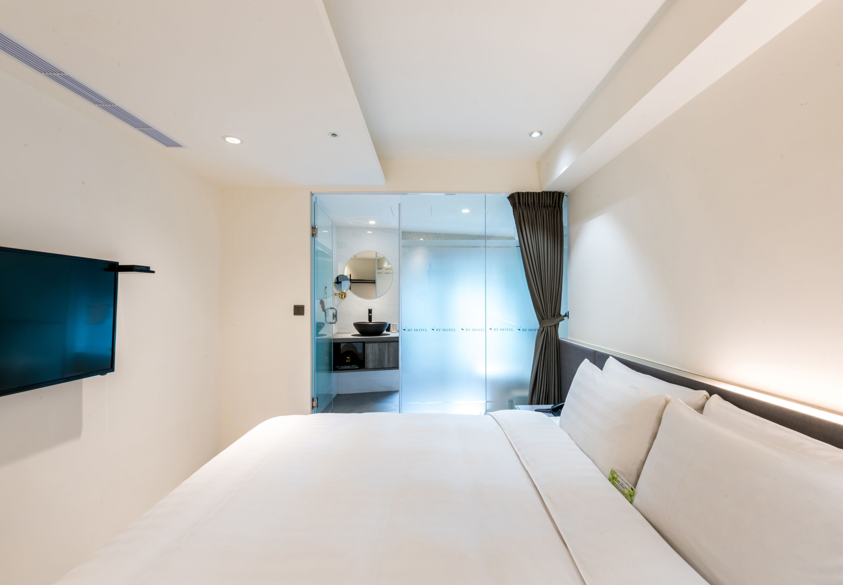 A modern hotel room with a large bed, a flat-screen TV mounted on the wall, a bathroom with a glass partition, and a view of the city.