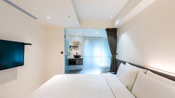 A modern hotel room with a large bed, a flat-screen TV mounted on the wall, a bathroom with a glass partition, and a view of the city.