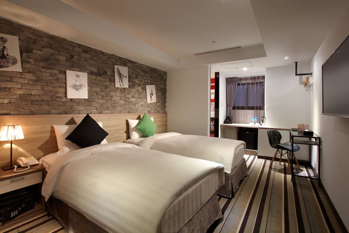 A hotel room with two beds, a desk, a TV, and artwork on the wall.