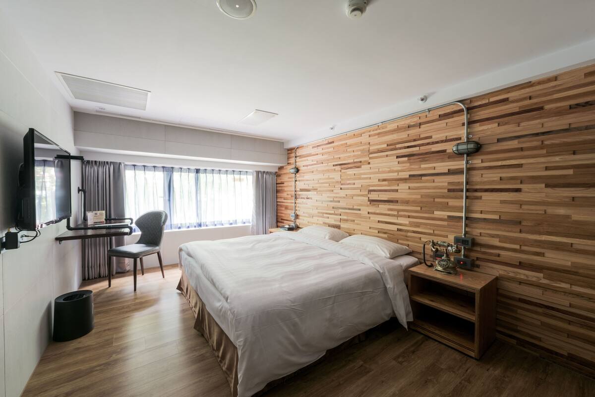 A modern hotel room with a wooden wall, a large bed, a desk with a chair, and a TV.