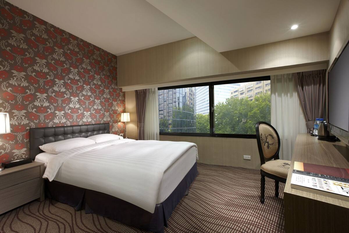 A hotel room with a large bed, a desk, a chair, a TV, and a window with a city view.