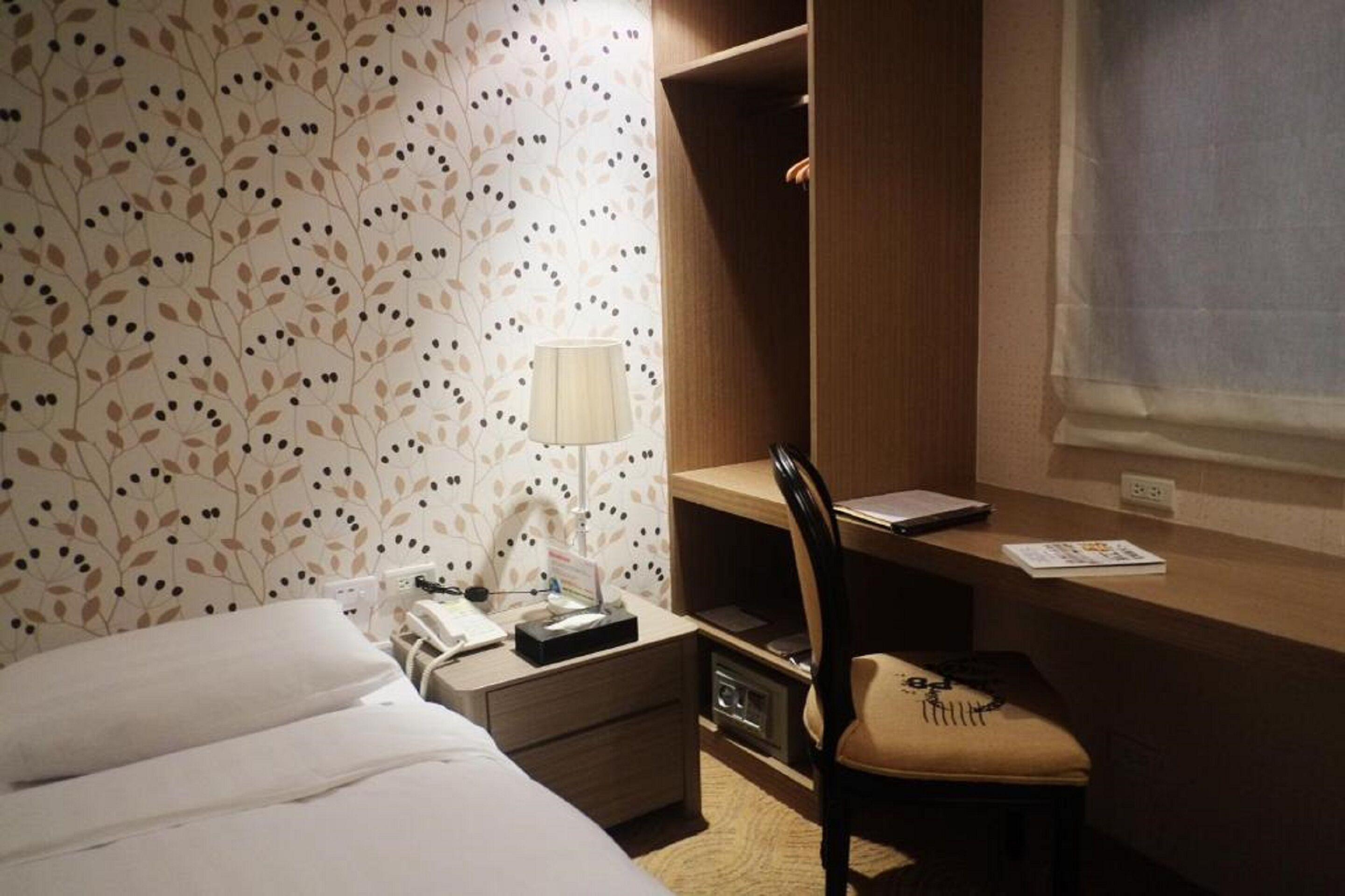 A hotel room with a bed, a desk, a chair, a lamp, and a window.