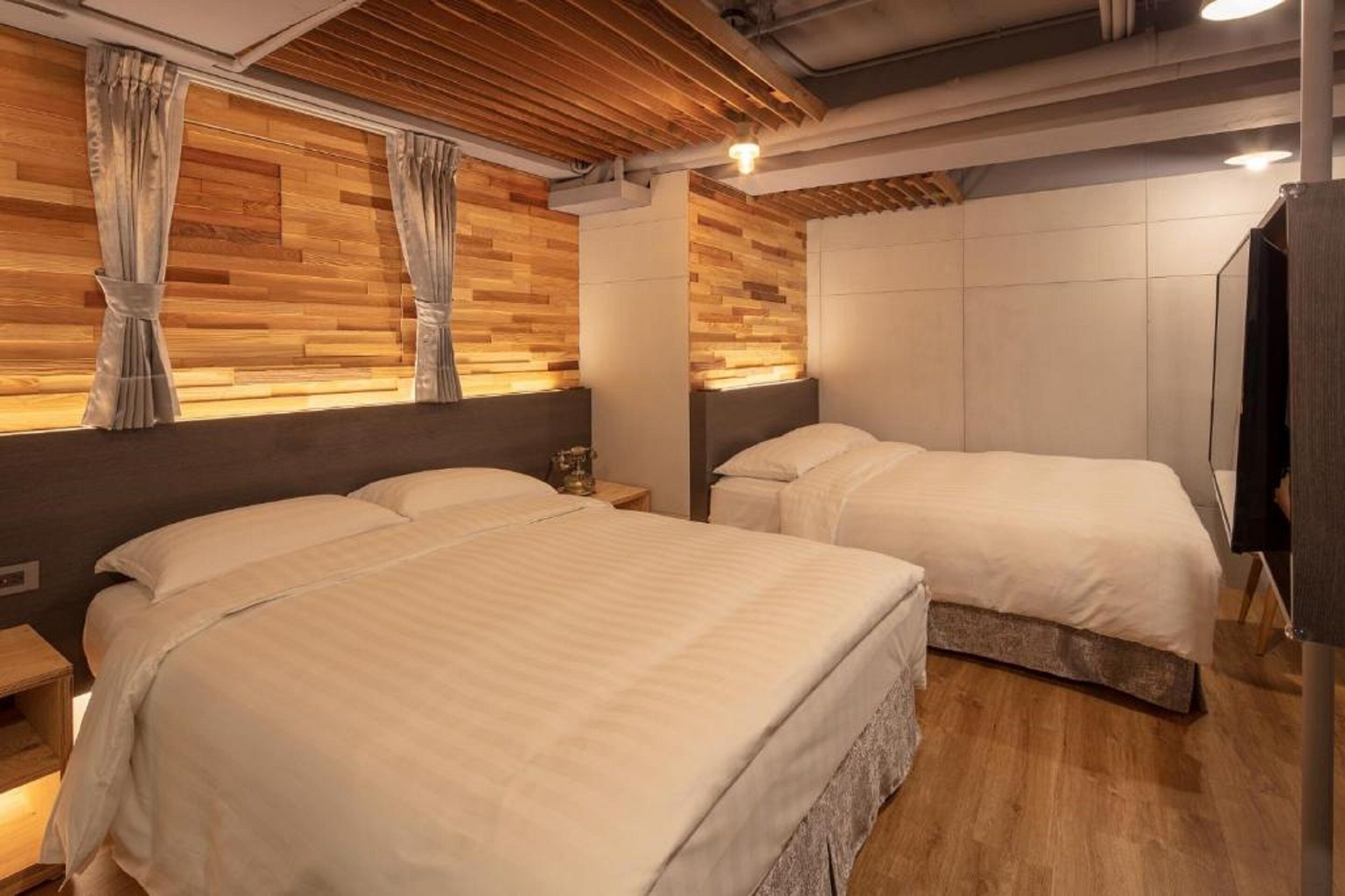 A modern hotel room with two beds, wooden panel walls, and a ceiling with exposed beams.