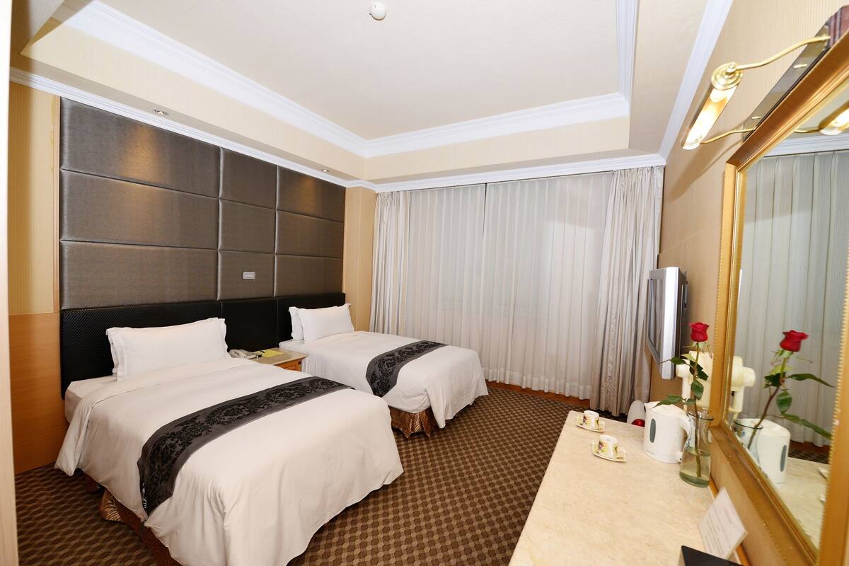 A hotel room with two beds, a large mirror, a bedside table with a lamp, and a small table with a vase of flowers.